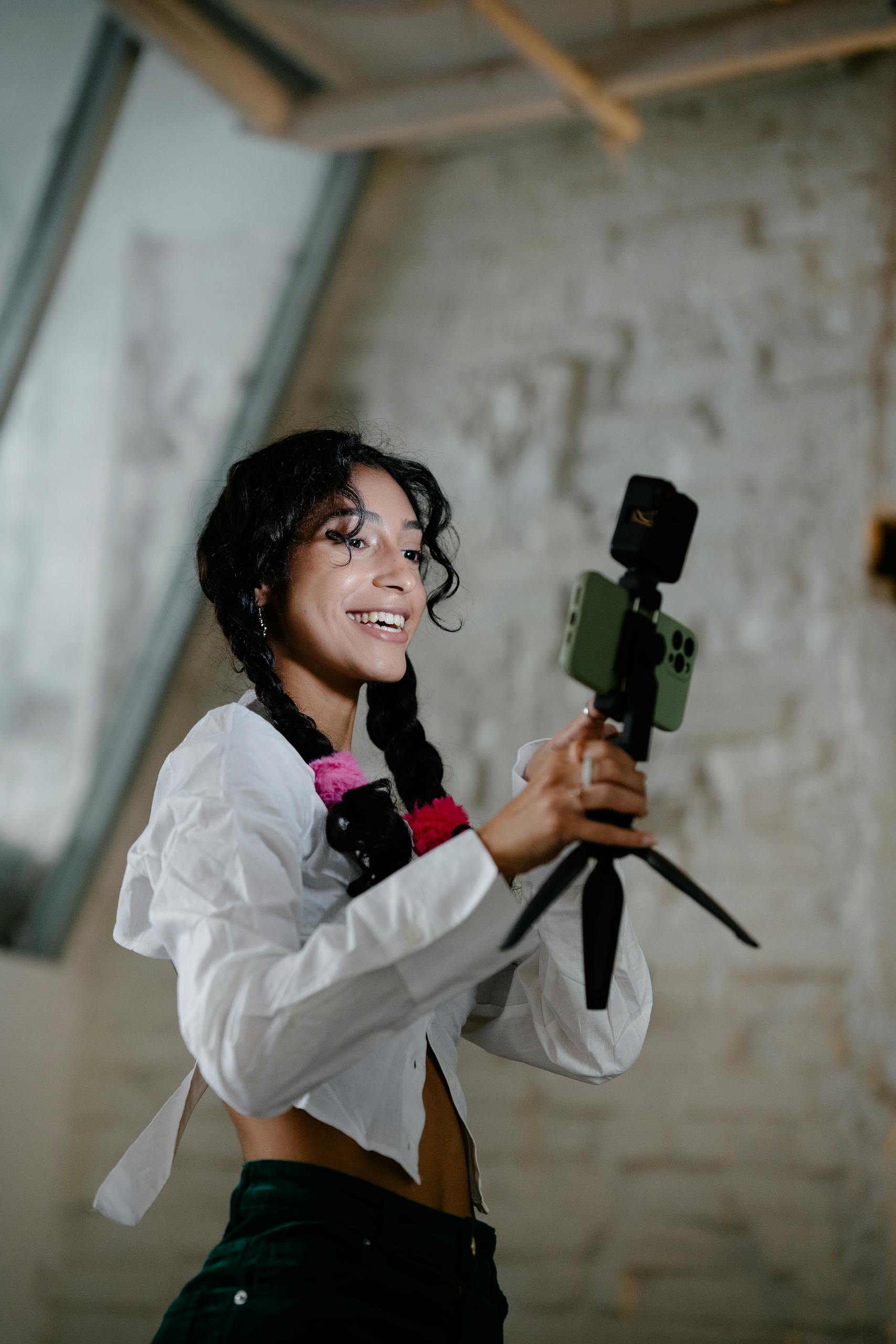 Happy woman with braids holds a smartphone mounted on a tripod indoors.